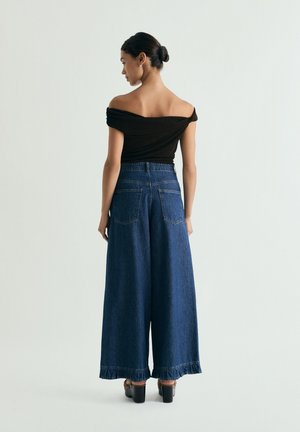 Mujer con el cabello recogido en un moño que lleva una blusa negra sin hombros, vaqueros anchos de mezclilla azul oscuro y zapatos negros de tacón, posando frente a un fondo gris liso.