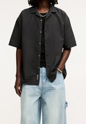 Person trägt ein schwarzes kurzärmeliges Hemd mit Knöpfen über einem schwarzen Top, hellblaue weite Jeans und eine silberne Kettenkette und Armband.