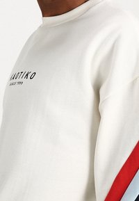 Felpa bianca con collare a costine, dotata di un logo nero e strisce rosse e blu lungo le maniche. Tessuto morbido.