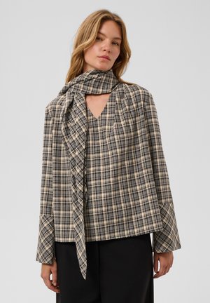 Femme portant un top à carreaux beige et noir avec une écharpe attachée drapée autour du cou et un pantalon noir, debout devant un fond uni.