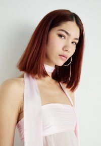 Abito di raso di un delicato rosa chiaro con scollo senza spalline e un lungo nastro al collo. La modella ha capelli lisci, di lunghezza spalla, di colore rossastro e indossa orecchini a cerchio.