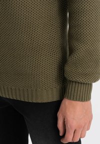 Olijfgroene gebreide trui met textuurpatroon, geribbelde manchetten en zoom, gedragen over zwarte jeans; close-up van de mouw en het polsgebied.