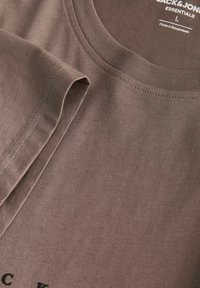 Gros plan d'un t-shirt en coton marron Jack & Jones Essentials montrant le col, la manche courte et l'étiquette de taille imprimée L fabriqué au Bangladesh.