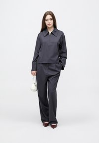 Νεαρή γυναίκα με σκούρο γκρι ζακέτα με φερμουάρ και παντελόνι με φαρδιά μπατζάκια, κρατώντας μια μικρή λευκή τσάντα, στέκεται μπροστά σε απλό ανοιχτόχρωμο φόντο.