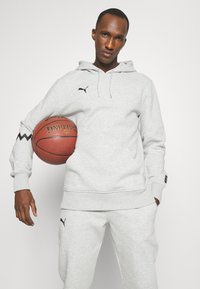 Šedá fleecová mikina s kangaroo vreckom, čiernym logom na hrudi a žltou detailom na rukávoch, v kombinácii so zodpovedajúcimi šedými teplákmi. Drží basketbal.