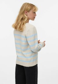 Pull beige clair avec des rayures horizontales bleu clair. Texture douce, col rond, poignets et ourlet côtelés. Porté avec un jean foncé.