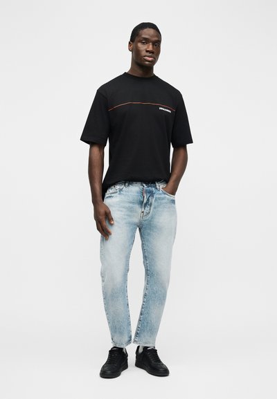 Άνδρας που στέκεται με το ένα χέρι στην τσέπη ανοιχτόχρωμων τζιν, φορώντας μαύρο T-shirt με πορτοκαλί γραμμή και μαύρα αθλητικά σε λευκό φόντο.