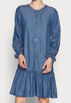 Femme portant une robe bleue à manches longues boutonnée avec des poignets froncés et un ourlet volanté, debout devant un fond uni.