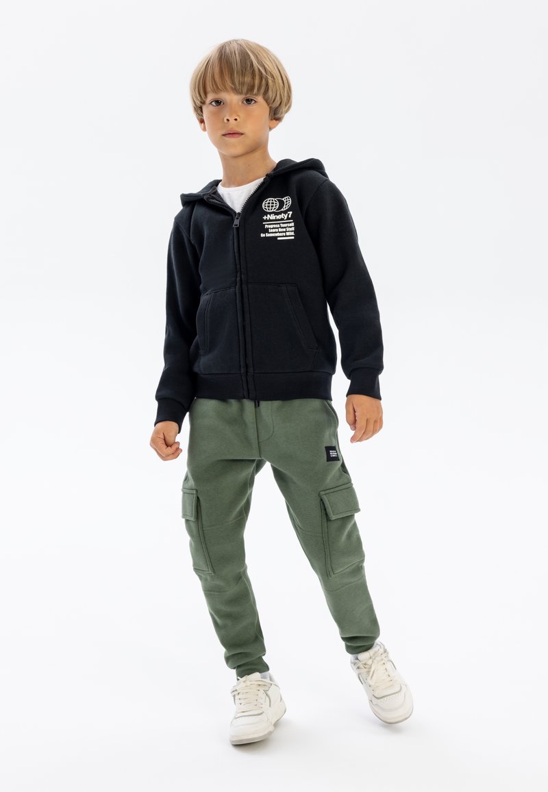 Musthave mustav kapuutsiga jakk, millel on ees taskud ja valge trükitud logo, koos roheliste cargo-pükstega, millel on küljetaskud ning valgete tossudega.