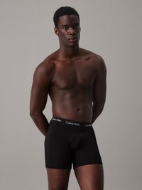 Crne pamučne bokserice s elastičnom trakom koja nosi Calvin Klein logo u bijeloj boji. Model se nalazi s pozadinom u neutralnim tonovima.