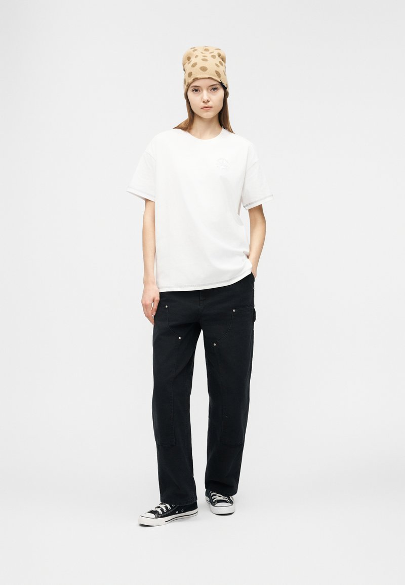 Giovane donna con berretto beige a pois, t-shirt bianca oversize, pantaloni neri larghi e sneaker nere basse, in piedi con una mano in tasca.