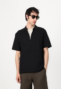 Polo shirt negru cu mâneci scurte și guler cu fermoar, asociat cu un tricou interior de culoare deschisă și pantaloni scurți de culoare verde măsliniu, având o textură a materialului fină.