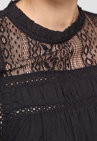 Schwarz Bluse mit hohem Kragen, Spitzenverzierungen mit Blumenmustern und strukturiertem Stoff. Rüschenbesatz entlang des Kragens fügt Detail hinzu.