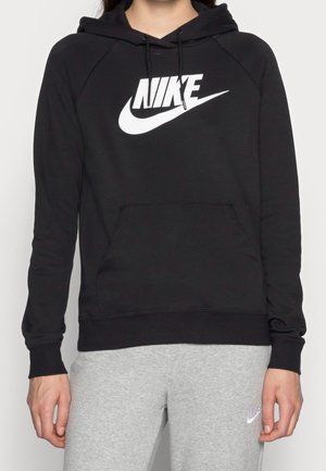 Person trägt schwarzen Nike-Hoodie mit weißem Logo und graue Nike-Sweatpants, steht vor einem schlichten hellen Hintergrund.