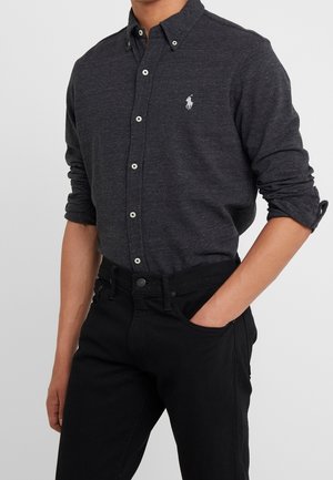 Mężczyzna ubrany w ciemnoszarą koszulę na guziki z długim rękawem, z podwiniętymi rękawami, oraz czarne spodnie, jedna ręka w kieszeni, na jednolitym tle.