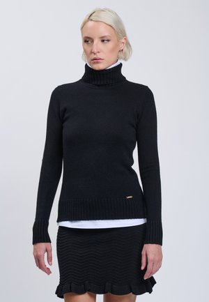 Femme blonde portant un pull à col roulé noir sur une chemise blanche et une jupe noire texturée avec un ourlet à volants, debout devant un fond uni.