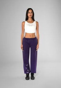 Witte cropped tanktop met een gestructureerd patroon; paarse wijde sweatpants met gedetailleerde accenten; zwarte sneakers.