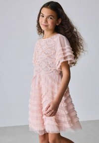 Fiatal lány hosszú, göndör hajjal, világos rózsaszín réteges ruhában, fodrokkal és gyöngydíszítésekkel, egyszínű háttér előtt állva.