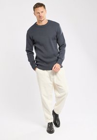 Marineblauer Strickpullover mit Rundhalsausschnitt, ausgestattet mit Schulterknöpfen, kombiniert mit creamfarbenen, weiten Hosen und schwarzen Stöckelschuhen vor einem schlichten Hintergrund.