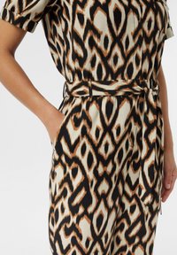 Wzorzysty kombinezon z beżowym tłem, ozdobiony czarnymi i brązowymi abstrakcyjnymi kształtami, z krótkimi rękawami, paskiem w talii oraz kieszeniami po bokach.