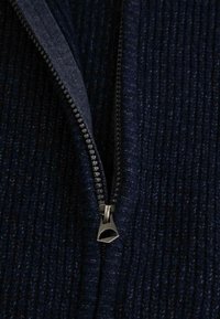 Granatowa kurtka z teksturowanej tkaniny z zamkiem błyskawicznym i metalowym uchwytem oraz prążkowanym wzorem. Skupienie na szczegółach zamka i jednolitości materiału.