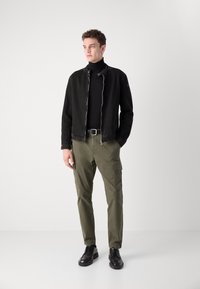 Chaqueta de mezclilla negra con cremallera plateada, suéter de cuello alto negro, pantalones cargo oliva con bolsillos y zapatos negros con cordones, de pie sobre un fondo neutro.