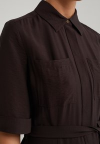 Rochie cămașă maro închis cu guler cu nasturi, mâneci scurte, două buzunare pe piept și talie legată, având un material neted și texturat.