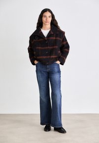 Cappotto di lana a quadri marrone e nero con ampio colletto e quattro bottoni, abbinato a jeans blu a campana e scarpe nere. Mani poggiate nelle tasche.