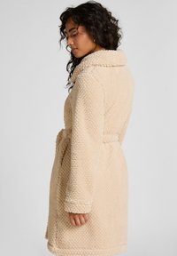 Beige Fleece-Rob mit einer strukturierten, flauschigen Oberfläche, großem Kragen und gebundenem Bund. Fällt bis zur Mitte des Oberschenkels und hat lange Ärmel.