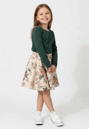 Uśmiechnięta dziewczyna w zielonym topie z długim rękawem, kwiecistej spódnicy i białych trampkach stoi ze splecionymi rękami na białym tle.