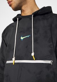 Homme portant un sweat à capuche noir avec des cordons blancs et une poche zippée horizontale blanche, avec un logo Nike bleu dégoulinant sur la poitrine.