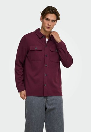 Jeune homme portant une veste bordeaux boutonnée avec poches poitrine et un pantalon gris, touchant son col sur fond blanc.