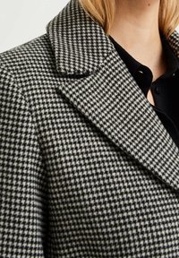 Cappotto a quadri grigio e nero con un grande colletto, caratterizzato da un tessuto testurizzato. Primo piano che mostra i dettagli del motivo e della lavorazione.