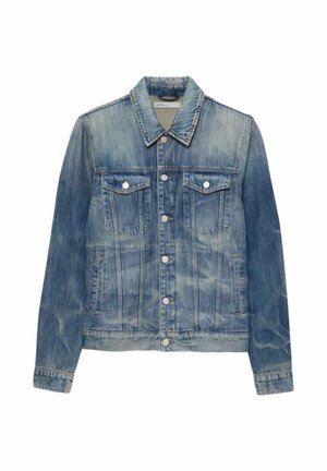 Jeune homme portant une veste en jean bleu délavée et un jean déchiré avec des bottes noires et des lunettes de soleil foncées, debout les mains dans les poches.
