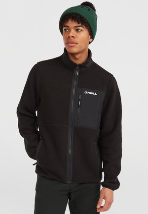 Young man wearing a black zip-up fleece jacket with a chest pocket and a green knit beanie, looking to the side.