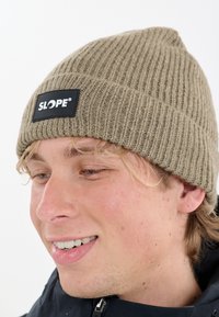 Nuori henkilö hymyilee päässään vaaleanruskea ribbikudottu pipo, jossa on musta "SLOPE" -logo-painatus, ja tummassa takissa, jossa on vetoketjukaulus.