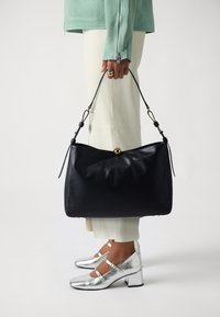Borsa a mano in pelle nera con forma morbida e arrotondata, accessori in oro e spallacci regolabili, tenuta da una persona che indossa scarpe argento e pantaloni chiari.