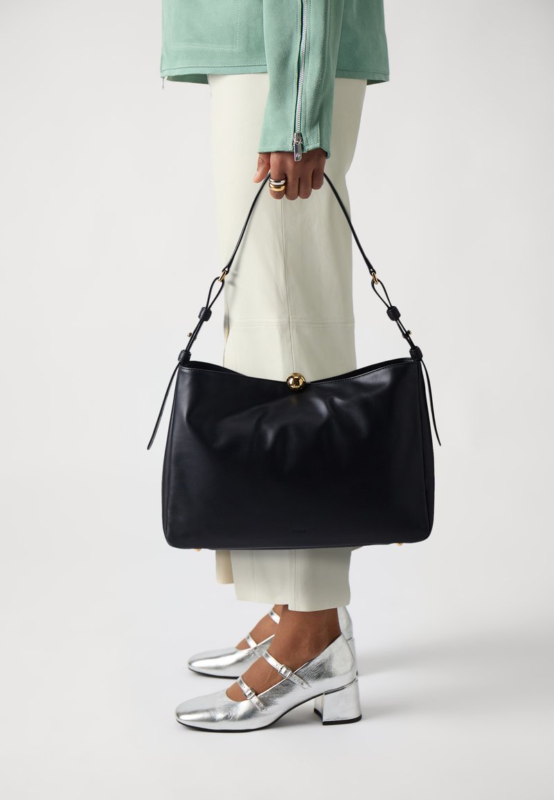 Borsa a mano in pelle nera con forma morbida e arrotondata, accessori in oro e spallacci regolabili, tenuta da una persona che indossa scarpe argento e pantaloni chiari.