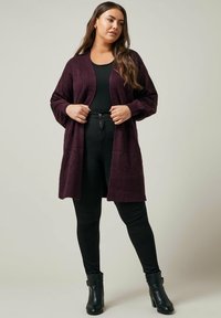Lång, öppen cardigan i djupt lila, tillverkad av strukturerat stickat tyg. Har breda ärmar och en avslappnad passform, parat med svarta byxor och stövlar.