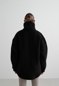 Personne avec les cheveux en chignon portant une veste polaire noire surdimensionnée avec un col montant, debout devant un fond gris clair uni.