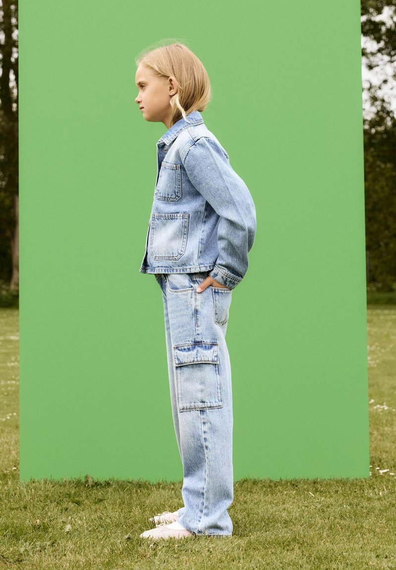 Giacca di jeans e pantaloni cargo di un blu chiaro. Giacca con tasche frontali; i pantaloni presentano una tasca cargo laterale. Sfondo verde, terreno erboso.