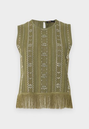 Femeie purtând o rochie fără mâneci cu model verde măsliniu și tiv cu franjuri, geantă neagră crossbody și sandale negre cu barete, stând pe fundal gri.