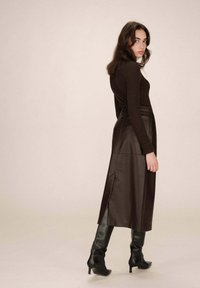 Top lungo a maniche ribbed marrone abbinato a una gonna in pelle marrone a vita alta con spacchi laterali, completato da stivali neri al ginocchio con tacco.