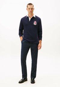 Marineblauwe polo met lange mouwen en een witte kraag, voorzien van een geborduurd embleem, gecombineerd met donkere gestructureerde broek en zwarte schoenen.