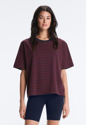 Donna che indossa una maglietta larga a righe rosse e blu navy e pantaloncini da biker blu navy, in piedi davanti a uno sfondo chiaro e uniforme.