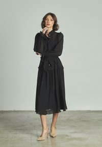 Vestido negro de manga larga con cintura con volantes y botones, hecho de una tela ligera, combinado con zapatos de tacón altos beige.