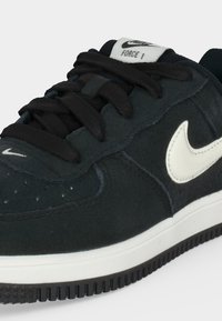 Μαύρο παπούτσι Nike Air Force 1 με λευκό λογότυπο swoosh, μαύρα κορδόνια και λευκή σόλα, εμφανιζόμενο σε κοντινή λήψη από μπροστινή πλευρική γωνία.