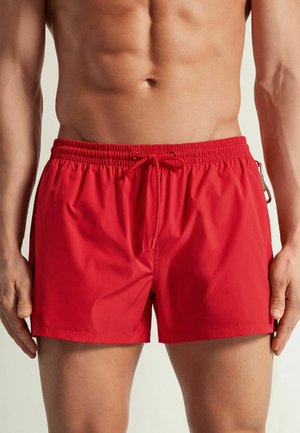 Gespiert mannelijk bovenlichaam met felrode zwemshorts met een elastische tailleband en een koordje aan de voorkant, staand met ontspannen armen langs het lichaam.