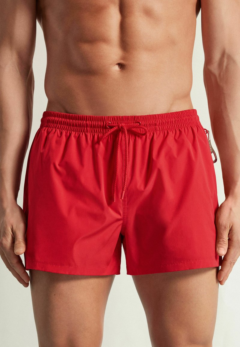 Torse masculin musclé portant un short de bain rouge vif avec une ceinture élastique et un cordon à l'avant, debout les bras détendus le long du corps.