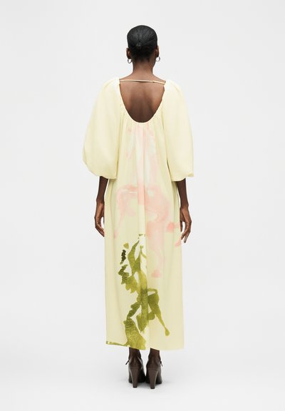 Γυναίκα με σκουρόχρωμο δέρμα και μαλλιά σε κότσο, φοράει μακρύ, ανοιχτό κίτρινο φόρεμα με φουσκωτά μανίκια, ανοιχτή πλάτη και λουλουδάτα σχέδια, στέκεται με την πλάτη προς τον θεατή.